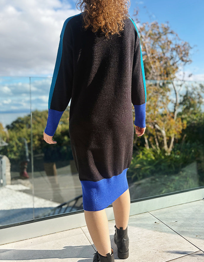 Woman wearing a black and blue colour block knitted sweater dress standing outdoors with trees and sky in the background