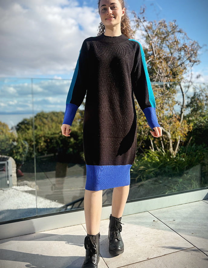 Woman wearing a black and blue colour block knitted sweater dress standing outdoors with trees and sky in the background