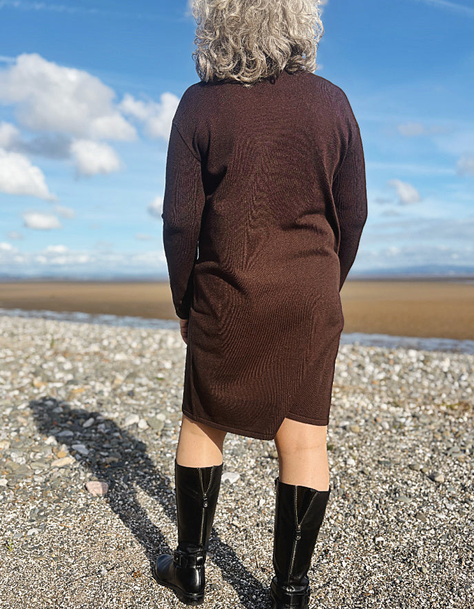 Person wearing a brown dress and black boots standing on a beach with a blue sky.