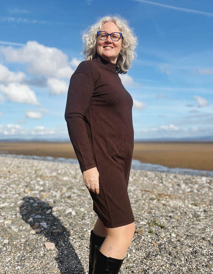 Woman in a brown dress standing on a beach with a blue sky and clouds in the background