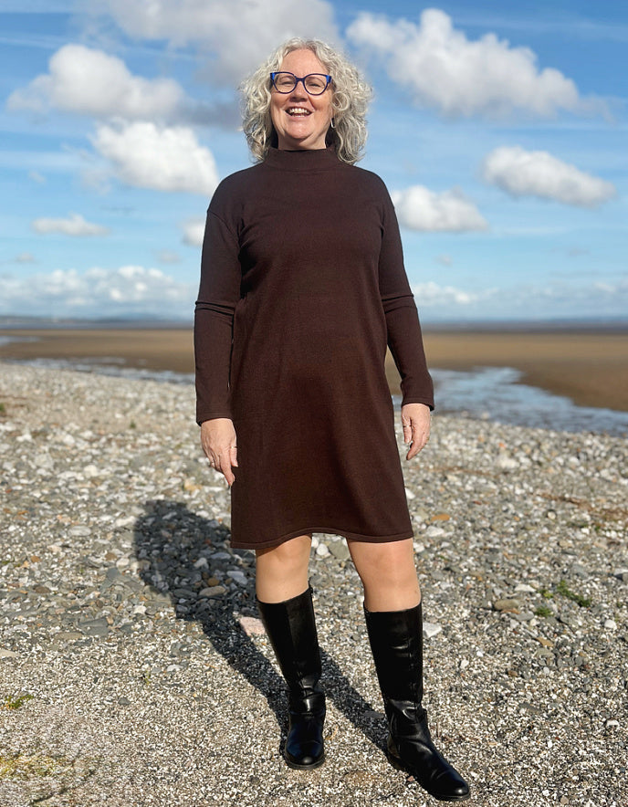Person wearing a brown dress and black boots standing on a pebbly beach with a blue sky.
