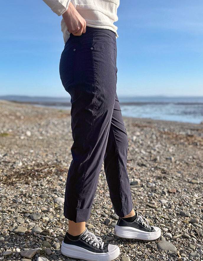 Person wearing navy pants and black sneakers standing on a pebbly beach with a clear blue sky.