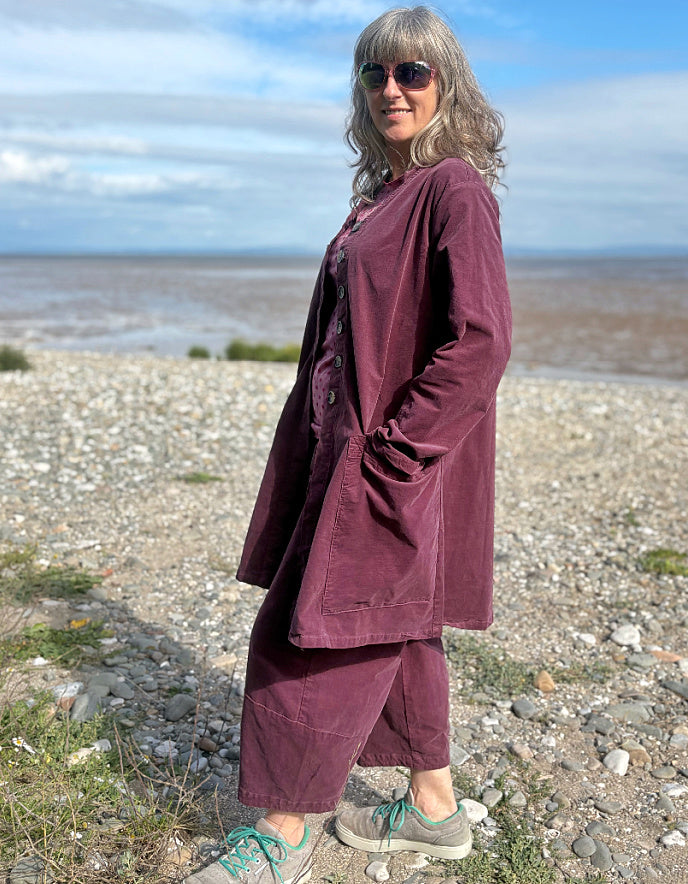 Woman in a burgundy plum corduroy duster coat and lantern shaped trousers standing on a pebbly beach with ocean in the background