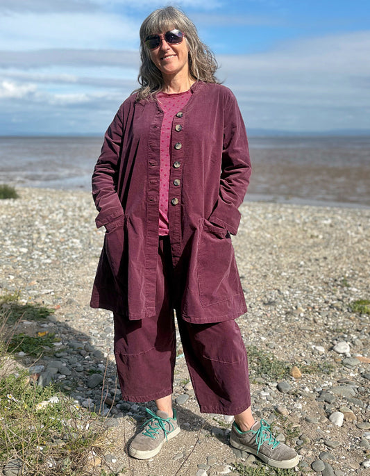 Woman in a burgundy plum corduroy duster coat and lantern shaped trousers standing on a pebbly beach with ocean in the background