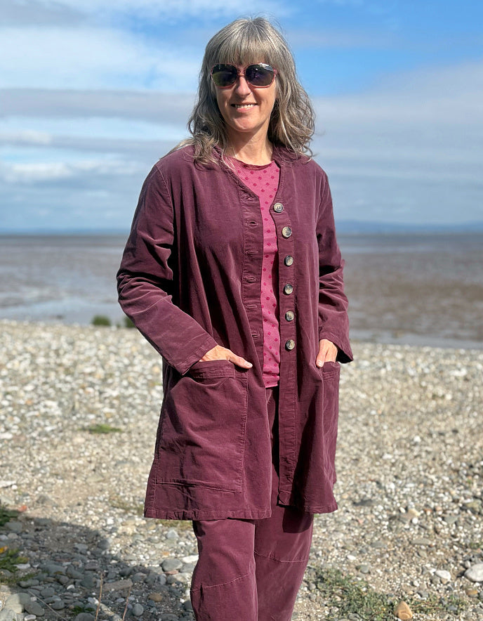 Woman in a burgundy plum corduroy duster coat and lantern shaped trousers standing on a pebbly beach with ocean in the background
