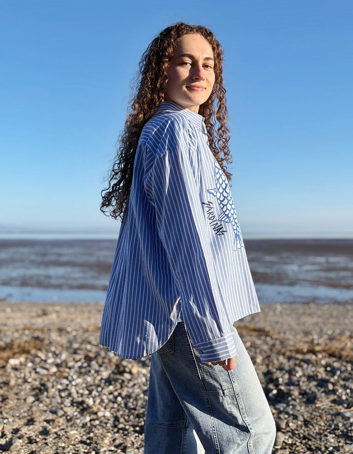 Person standing on a pebbly beach with ocean view wearing a blue and white striped shirt.