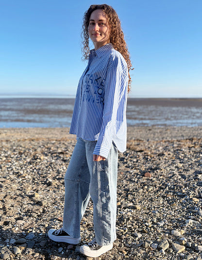 Person standing on a rocky beach wearing a blue and white striped shirt and jeans.