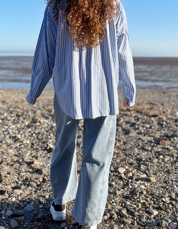 Person wearing a blue and white striped shirt and jeans standing on a pebbly beach.