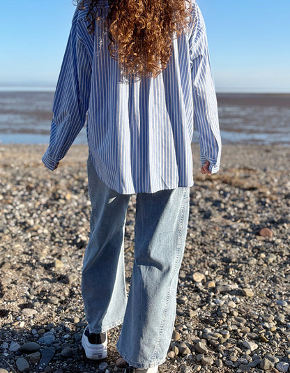 Person wearing a blue and white striped shirt and jeans standing on a pebbly beach.
