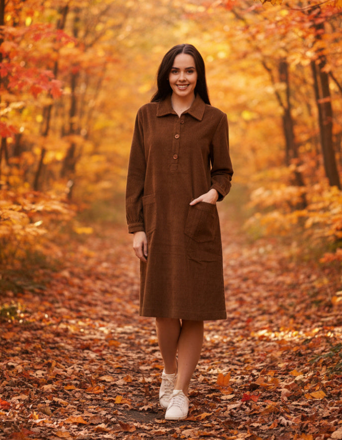 Woman in a brown cord dress standing in an autumn forest with orange and yellow leaves.
