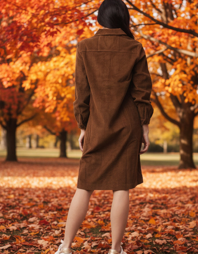 woman wearing a brown cord dress standing in an autumn park with orange leaves.