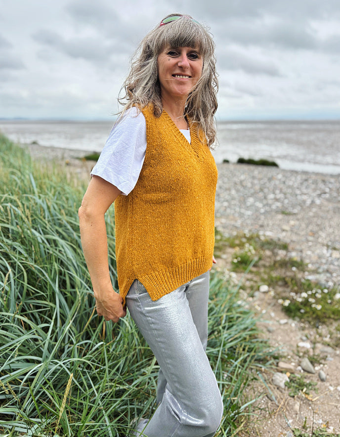 Woman wearing a yellow knitted vest and white shirt standing on a beach.