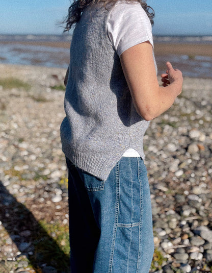 A person standing on a rocky beach wearing a gray ribbed knit sleeveless vest over a white T-shirt and blue jeans, photographed from the side in natural outdoor light.
