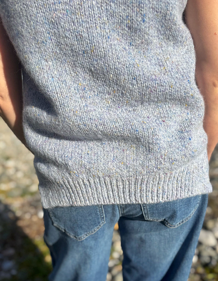 Close-up of a person wearing a light gray knit sweater vest with multicolored flecks and blue jeans, standing outdoors.