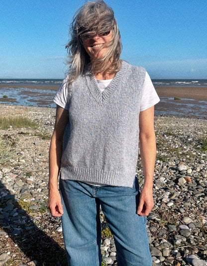 A woman standing on a rocky beach wearing a light gray knit V-neck sweater vest over a white t-shirt and blue jeans.