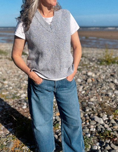 A woman standing on a rocky beach wearing a light gray ribbed knit sweater vest over a white t-shirt and blue jeans.