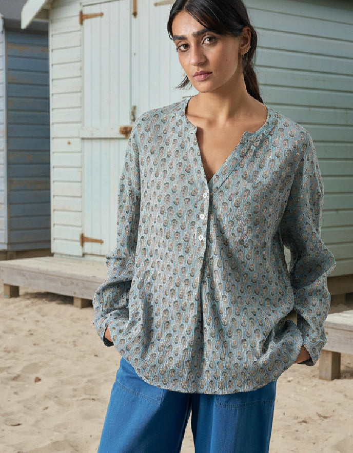Woman wearing a patterned blouse and blue pants on a sandy beach with wooden cabins in the background.