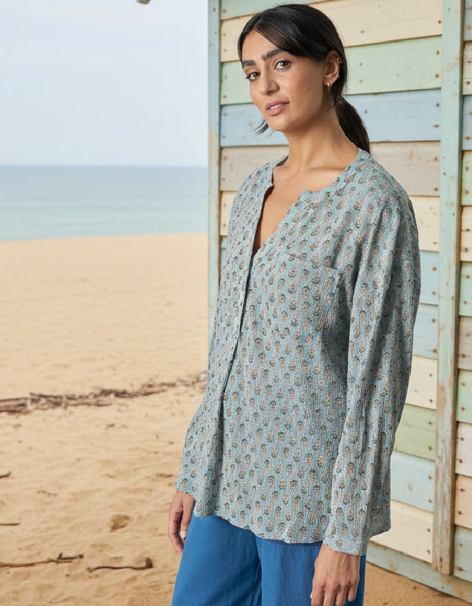 Woman wearing a light blue patterned shirt and blue pants standing on a beach.