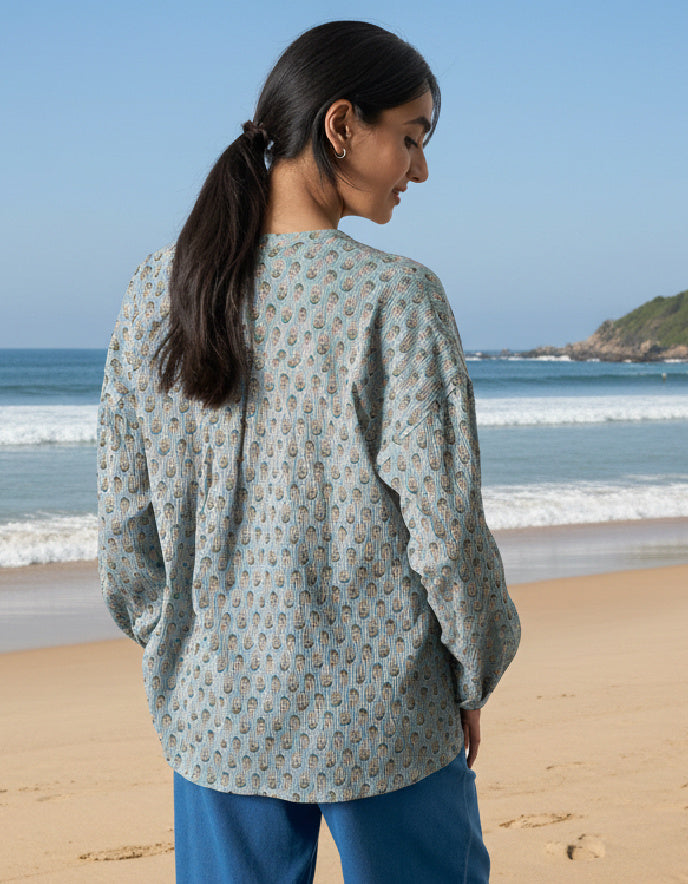 Woman standing on a beach wearing a patterned shirt and blue pants, looking out towards the ocean.
