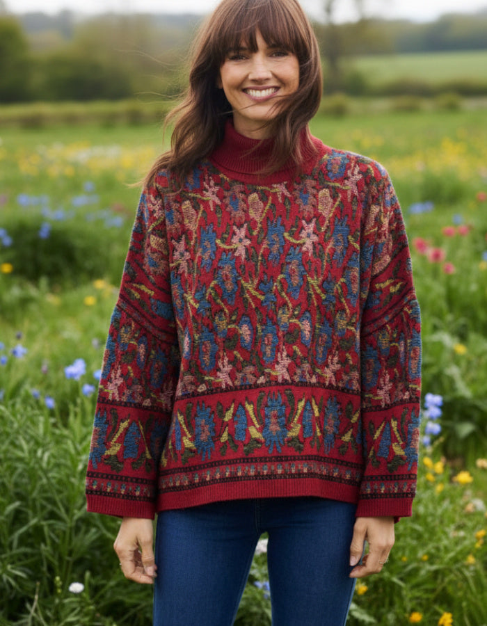 A woman wearing a red turtleneck sweater with an intricate multicolor floral and geometric pattern, paired with blue jeans, standing in a lush green field of wildflowers.