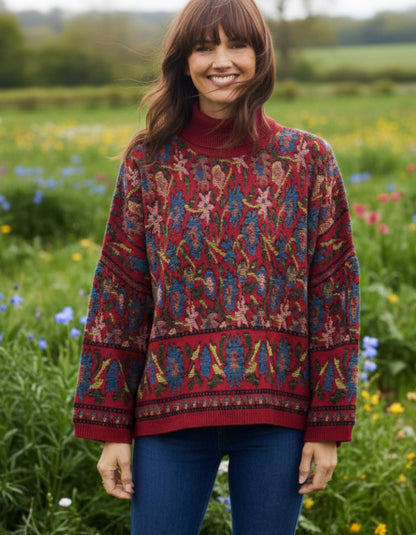 A woman wearing a red turtleneck sweater with an intricate multicolor floral and geometric pattern, paired with blue jeans, standing in a lush green field of wildflowers.