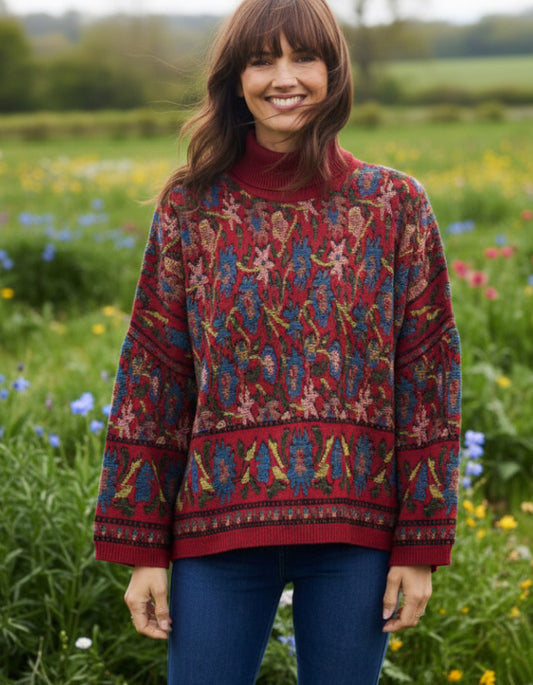 A woman wearing a red turtleneck sweater with an intricate multicolor floral and geometric pattern, paired with blue jeans, standing in a lush green field of wildflowers.