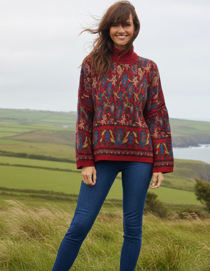 A woman wearing a long-sleeve turtleneck sweater with a complex multicolor geometric and tribal pattern in red, blue, yellow, and green, paired with blue jeans, standing outdoors in a grassy field with hills and water in the background.