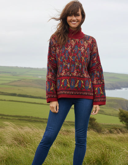A woman wearing a long-sleeve turtleneck sweater with a complex multicolor geometric and tribal pattern in red, blue, yellow, and green, paired with blue jeans, standing outdoors in a grassy field with hills and water in the background.