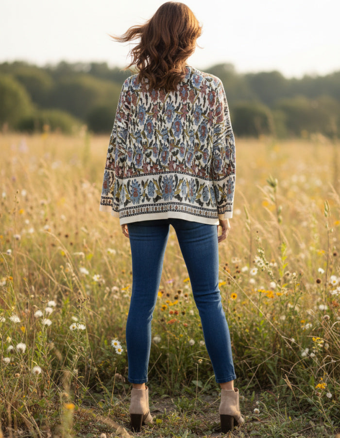A woman stands in a sunlit grassy field with wildflowers, photographed from behind. She is wearing a loose, long-sleeve patterned cardigan or sweater in beige, blue, and brown tones, paired with blue skinny jeans and tan ankle boots.