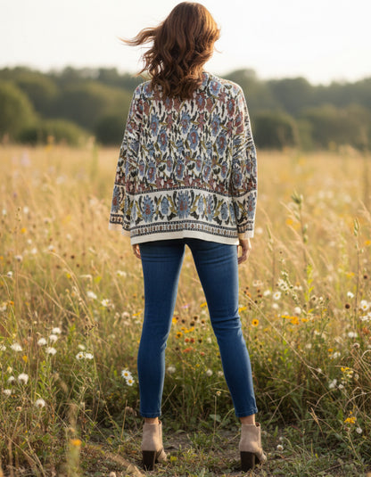 A woman stands in a sunlit grassy field with wildflowers, photographed from behind. She is wearing a loose, long-sleeve patterned cardigan or sweater in beige, blue, and brown tones, paired with blue skinny jeans and tan ankle boots.