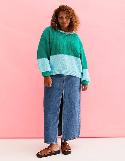 A woman modeling a color-block knit sweater in green and light blue, paired with a blue denim midi skirt and brown sandals, set against a solid pink studio background.
