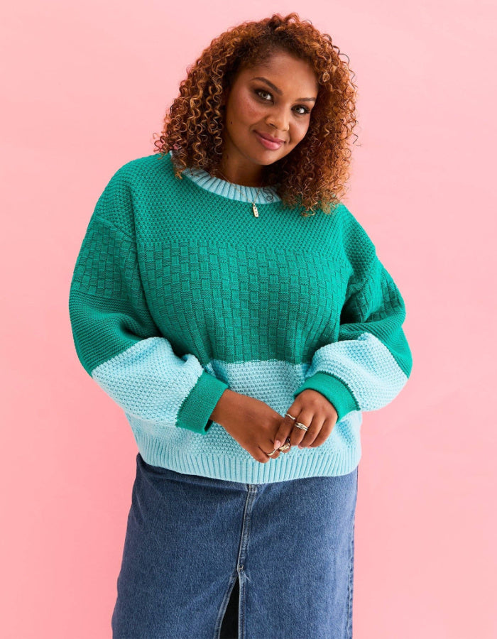 A woman with curly brown hair wearing a green and light blue color-blocked knit sweater and a blue denim skirt, posing against a solid pink background.