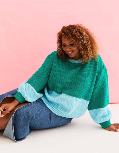 A woman with curly light brown hair sits on a white floor against a solid pink background, wearing a color-blocked knit sweater with a green body, light blue ribbed hem and cuffs, and blue jeans.