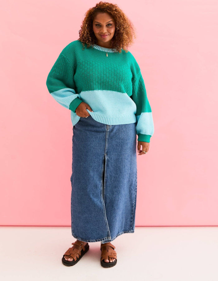 A woman modeling a colorblock knit sweater in green and light blue, paired with a long blue denim skirt and brown sandals, set against a solid pink studio background.
