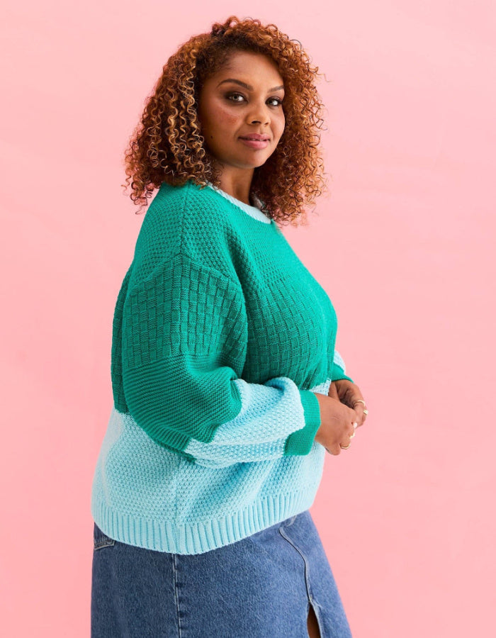 A woman with curly brown hair wearing a two-tone green and light blue knit sweater, paired with a denim skirt, posing against a solid pink background.