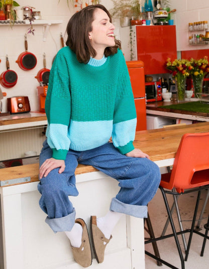 A woman wearing a color-block knit sweater in teal green and light blue, paired with cuffed blue jeans, white socks, and tan slip-on shoes, sitting on a kitchen counter in a retro-styled kitchen.