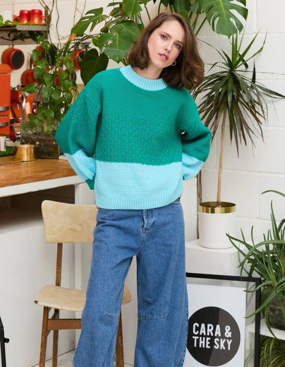 A woman wearing a green and light blue color-block knit sweater and wide-leg blue jeans, standing in a styled indoor setting with plants, a wooden chair, and a sign that reads 'CARA & THE SKY'.