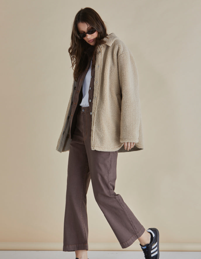 Woman wearing a beige sherpa pile fleece shirt style coat and brown pants against a beige background