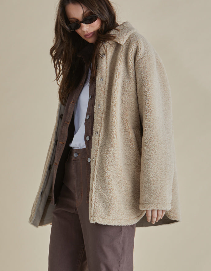 Woman wearing a beige sherpa pile fleece shirt style coat and brown pants against a beige background