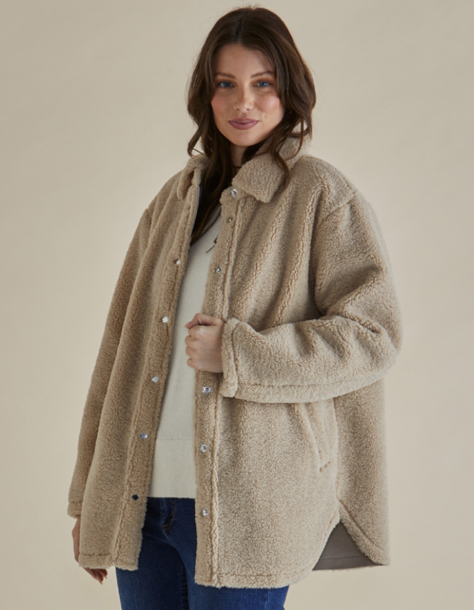 Woman wearing a beige sherpa pile fleece shirt style coat and brown pants against a beige background