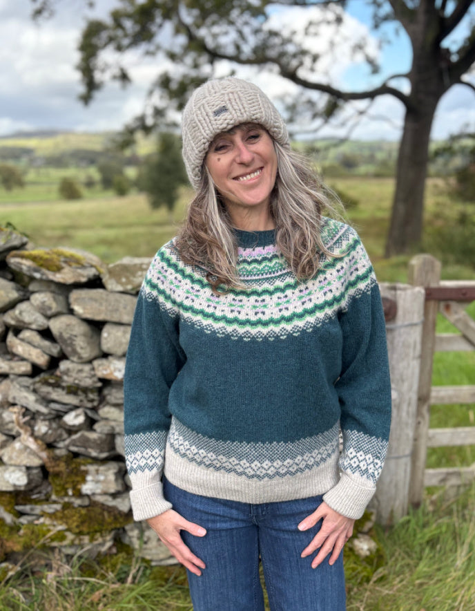 Woman wearing a patterned sweater and beanie standing in a rural landscape with a stone wall and wooden gate.
