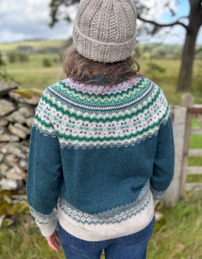 Person wearing a blue knitted sweater with a patterned design, standing outdoors in a natural setting.