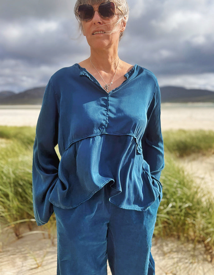 A woman wearing a matching blue long-sleeve top and pants set stands outdoors on a sandy beach with dune grass and a cloudy sky in the background.