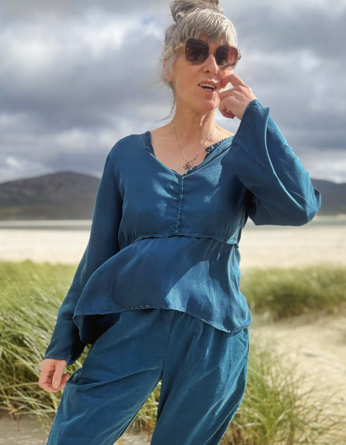 A woman wearing a matching blue long-sleeve top and pants set, styled with sunglasses and a necklace, standing outdoors in a grassy field with mountains and a cloudy sky in the background.