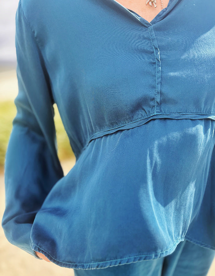 A close-up of a person wearing a blue long-sleeve top with a V-neck and a gathered peplum-style hem, photographed outdoors with a softly blurred background.