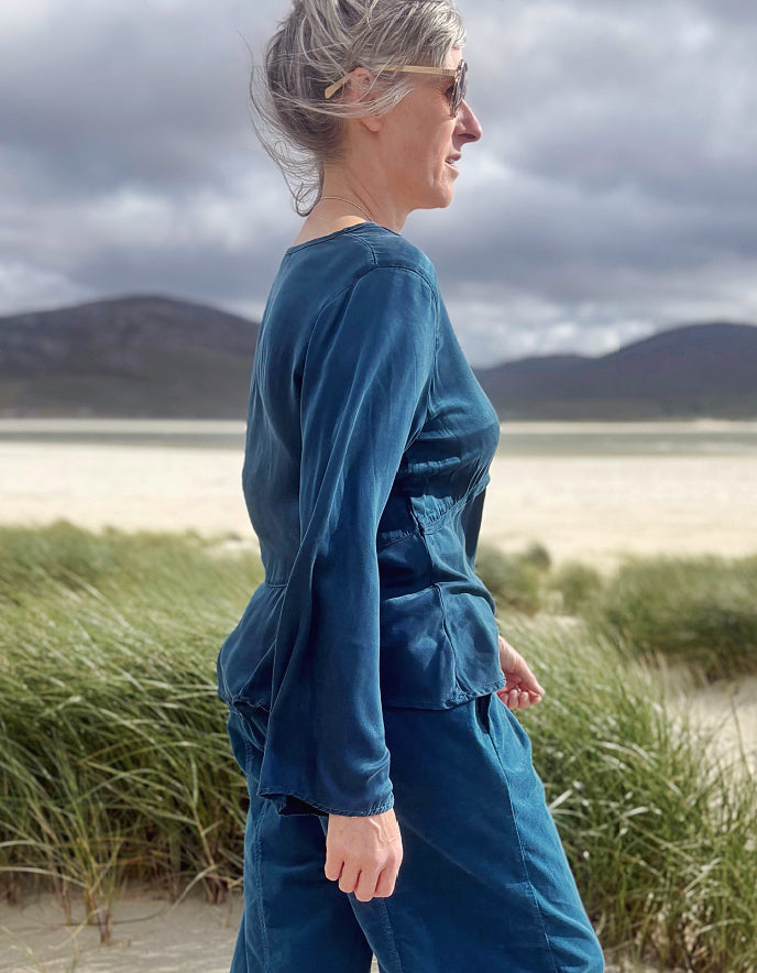 A woman wearing a matching teal blue long-sleeve top and trousers stands in profile on a beach dune with grass and distant hills under a cloudy sky.