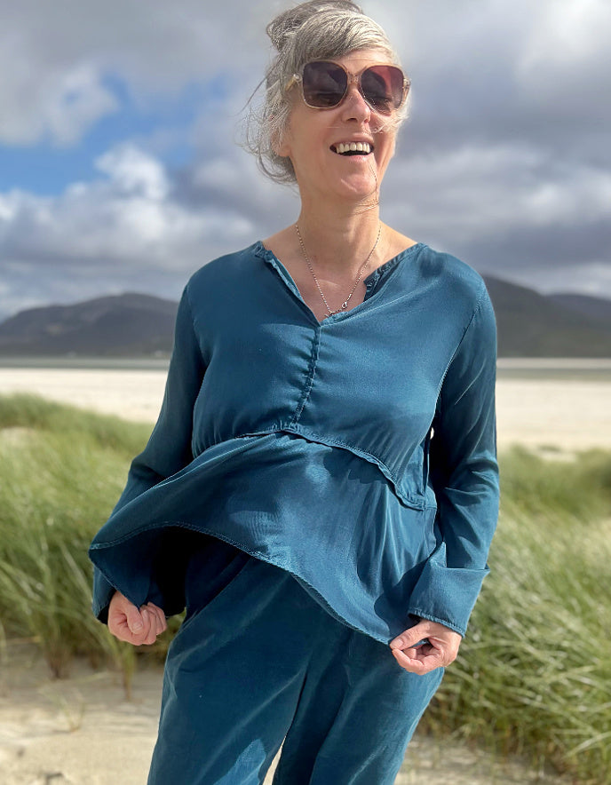 A woman with gray hair and sunglasses stands outdoors on a sandy beach path wearing a teal blue long-sleeve top and matching pants, with dune grass and mountains in the background.