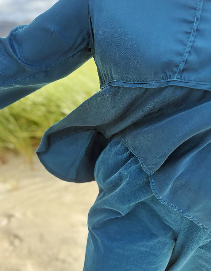 A close-up outdoor shot of a person wearing a textured blue long-sleeve top or jumpsuit, with the fabric gathered at the waist and a blurred beach or dune background.