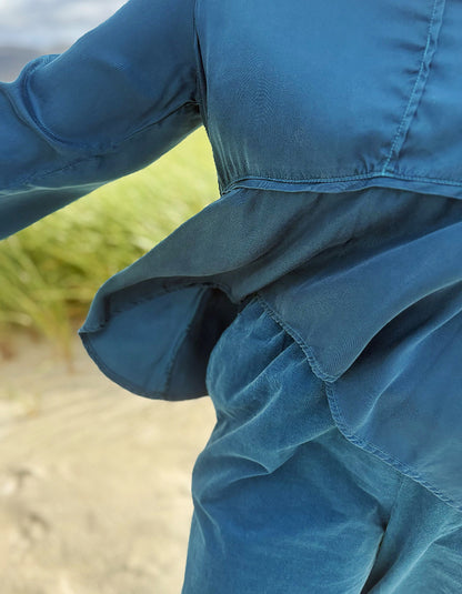 A close-up outdoor shot of a person wearing a textured blue long-sleeve top or jumpsuit, with the fabric gathered at the waist and a blurred beach or dune background.