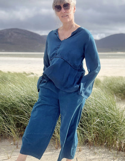 A woman wearing a matching blue long-sleeve top and wide-leg pants set stands outdoors on a sandy beach with tall grasses and mountains in the background.